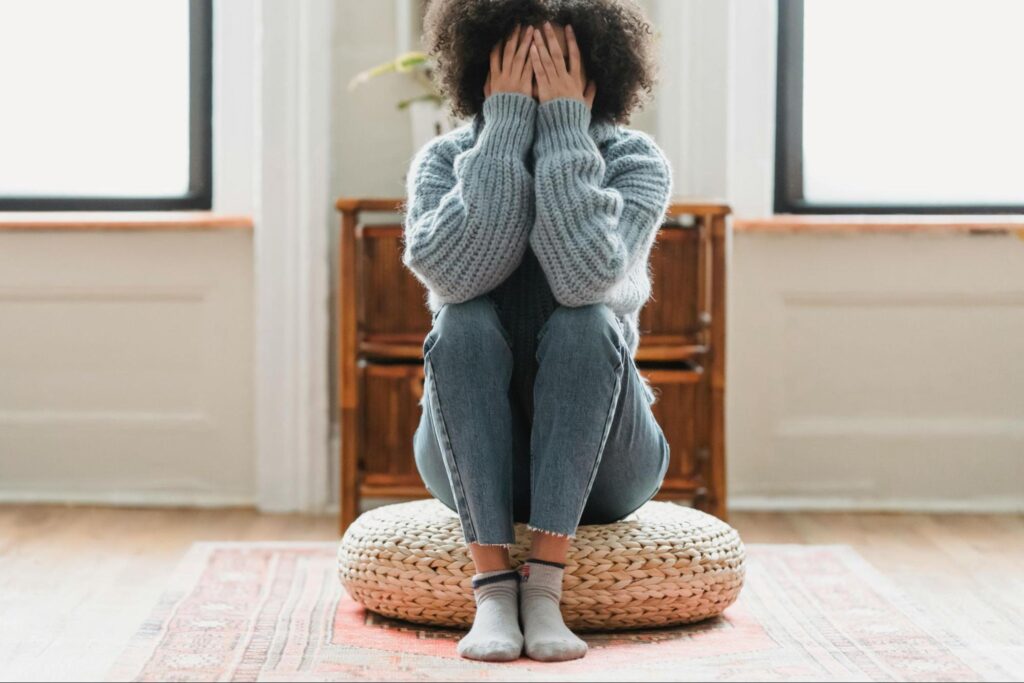 Young person covering face with hands, sitting on a round cushion in a bright room